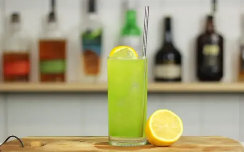 Zielony, orzeźwiający drink Tokyo Iced Tea z plasterkiem cytryny i metalową słomką, na tle półki z butelkami alkoholu.