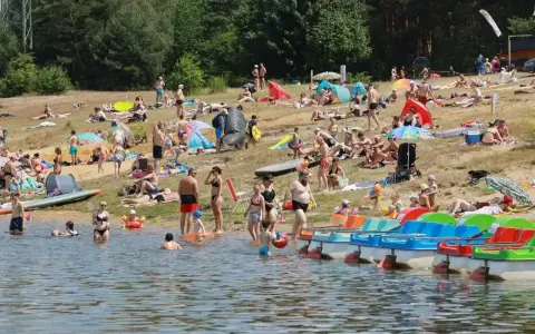 Tłumy ludzi wypoczywają na plaży nad jeziorem Chechło. Woda jest pełna kąpiących się, a na brzegu opalają się plażowicze.