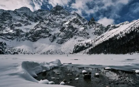 Zamarznięte Morskie Oko, otoczone ośnieżonymi szczytami Tatr. W oddali widać ludzi spacerujących po lodzie, a na pierwszym planie płynie strumyk.