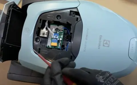 En person med svarta handskar skruvar på ett kretskort inuti en ljusblå Electrolux dammsugare, märkt "PURE D8.2 SILENCE".