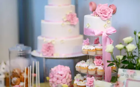 Eleganckie, białe torty weselne i babeczki ozdobione różowymi kwiatami i wstążkami. Idealne na słodki stół, inspiracja jakie ciasta na wesele.