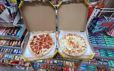 Dwie pizze w pudełkach, jedna z salami, druga serowa. Obok półki ze słodyczami i napojami w sklepie Żabka.