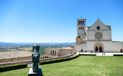 Statua equestre domina il prato verde di Assisi. Cosa vedere: la Basilica di San Francesco, con la sua imponente facciata e campanile, si staglia contro il cielo azzurro.