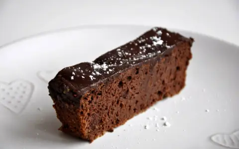 Wilgotny brownie z czerwonej fasoli, polany czekoladą i posypany cukrem pudrem, na białym talerzu z serduszkami.