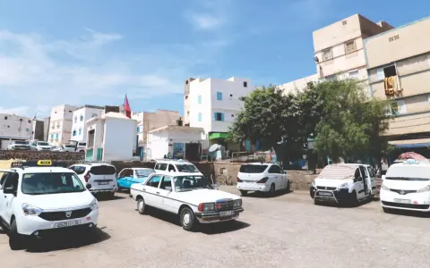 Taxis warten auf Fahrgäste in einer sonnigen Stadt in Marokko. Weiße Gebäude und Bäume säumen die Straße.
