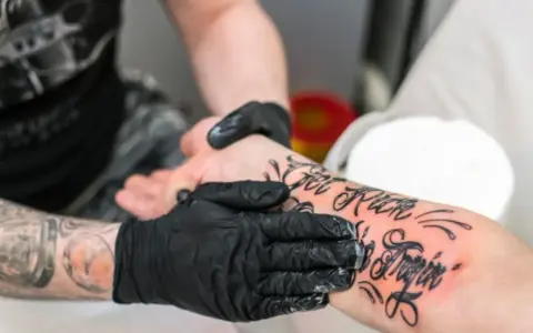 Ręce w czarnych rękawiczkach smarują świeży tatuaż na przedramieniu. Pytanie: kiedy zacząć smarować tatuaż?