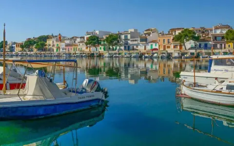Boote im Hafen von Portocolom, Mallorca, spiegeln sich im ruhigen, blauen Wasser vor bunten Häusern.