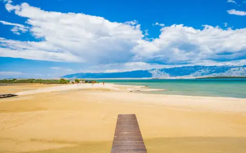Drewniany pomost prowadzi na piaszczystą plażę Ninskiej laguny. W tle turkusowe morze i góry.