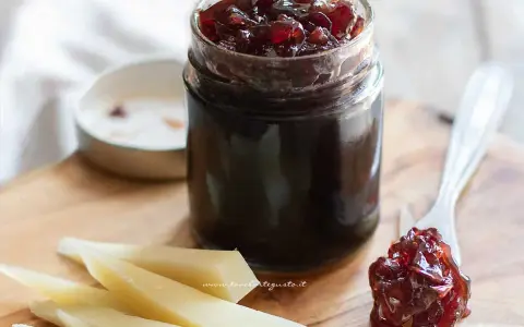 Un vasetto di marmellata di cipolle scura, con un cucchiaino che ne raccoglie una porzione, accanto a fette di formaggio stagionato.