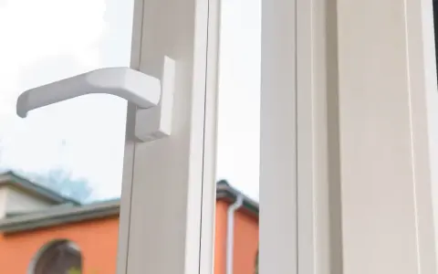Poignée blanche d'une fenêtre, révélant le mécanisme cremone porte fenetre. Dehors, des maisons et des arbres verts.