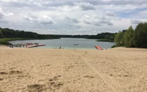Piaskowa plaża nad jeziorem, z pomostami i łódkami. W oddali las, chmury na niebie. Idealne miejsce na wypoczynek nad wodą.