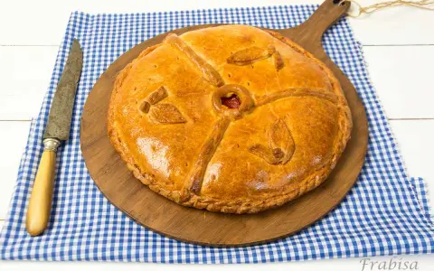 Empanada dorada y decorada sobre tabla de madera, con cuchillo y mantel a cuadros.