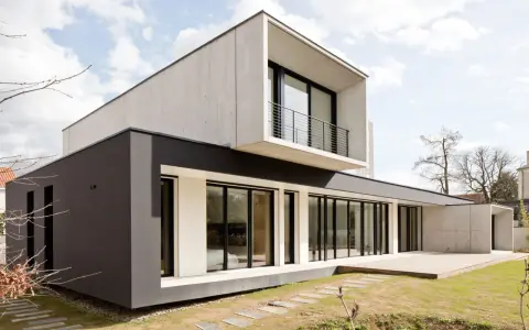Une maison moderne aux lignes épurées, avec des blocs de béton et de crépi sombre, dotée de grandes baies vitrées et d'une terrasse en bois.