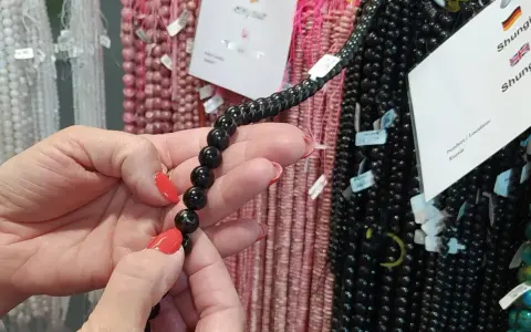 Manos con uñas rojas sostienen un collar de cuentas negras de shungita. En el fondo, se ven hilos de cuentas rosas y blancas.