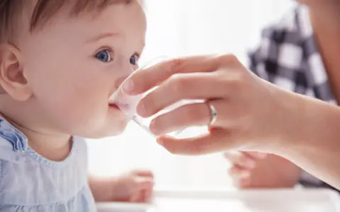 Ein Baby trinkt vorsichtig Wasser aus einem Becher, gehalten von einer Hand. Die Frage "ab wann Baby Wasser geben?" wird hier visuell beantwortet.