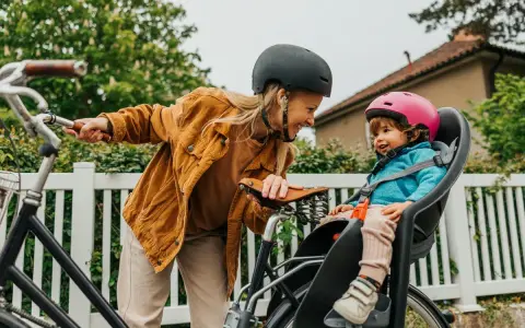 En kvinna med hjälm ler mot ett litet barn i en cykelbarnstol. De är redo att cykla med bebis i det vackra vädret.