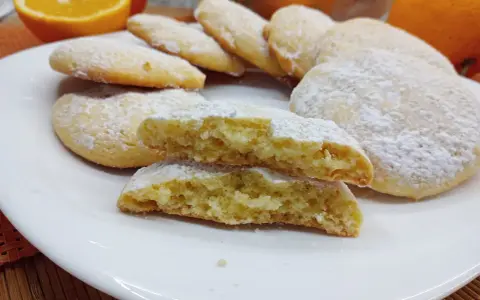 Biscotti morbidi all'arancia spolverati di zucchero a velo su un piatto bianco, con arance fresche sullo sfondo.