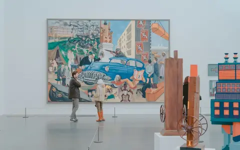 Two people observe a large, colorful mural depicting a chaotic scene with a blue car, factories, and people, offering a glimpse into the art market definition of diverse artistic expression.