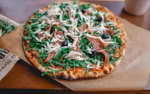 Pyszna pizza prosciutto z rukolą, oliwkami i parmezanem, podana na papierze do pieczenia.