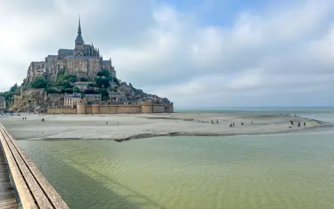 St. Michel, eine beeindruckende Abtei auf einer Felseninsel, ragt über einer sandigen Bucht auf. Menschen spazieren am Strand, während das Wasser sanft ansteigt.