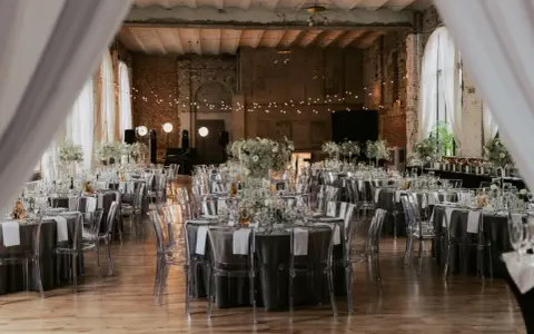 Eleganckie wesele w industrialnym stylu. Stoły nakryte szarymi obrusami, ozdobione białymi kwiatami i transparentnymi krzesłami. Girlandy świateł tworzą magiczny nastrój.