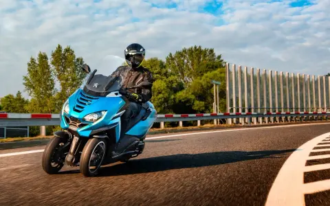 Niebieski Peugeot Metropolis 400 z kierowcą w kasku pędzi po zakręcie drogi, otoczony zielenią i błękitnym niebem.