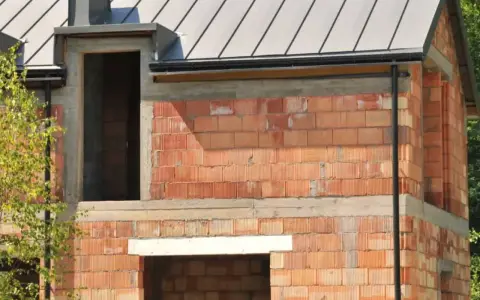 Budynek w budowie z cegły, widoczne okna i drzwi, dach pokryty blachą. Widać cofnięte nadproże do rolet.