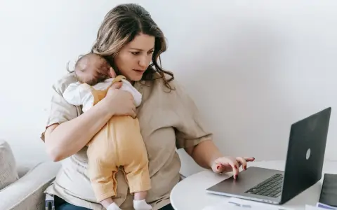 Zmęczona kobieta z dzieckiem na rękach pracuje przy laptopie. To obraz zespołu stresu opiekuna, gdzie obowiązki domowe i zawodowe nakładają się na siebie.