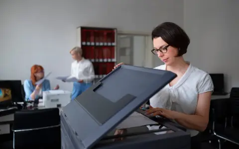 Frau mit Brille bedient einen Drucker, vielleicht um einen Epson Drucker zu installieren. Im Hintergrund arbeiten Kolleginnen.