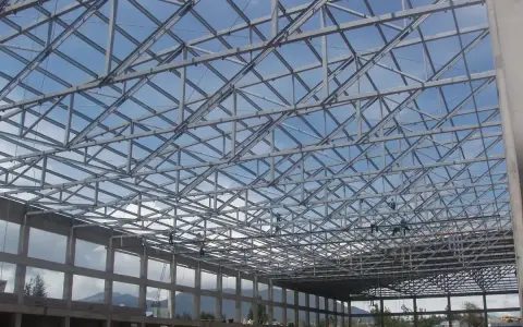 Estructura metálica de una **cercha Pratt** en construcción, con operarios trabajando en el techo bajo un cielo azul.