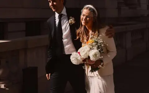 Novia sonriente con velo y ramo, luciendo una chaqueta novia boho sobre su vestido. El novio, con traje oscuro, la abraza.