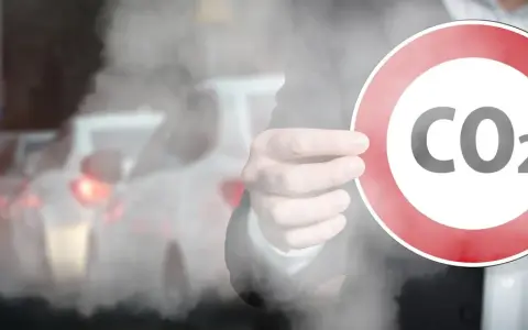 Dłoń trzyma znak zakazu z napisem CO2 na tle dymiących samochodów w korku.