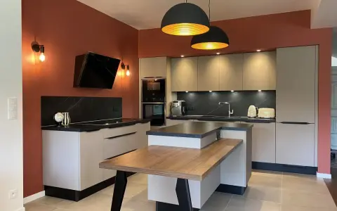 Cuisine moderne aux murs terracotta, avec îlot central en bois et mobilier gris clair. Éclairage chaleureux.