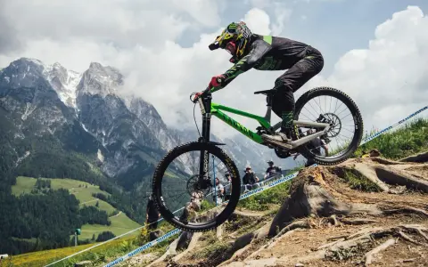 Troy Brosnan in mid-air on a green downhill bike, with a dramatic mountain backdrop.