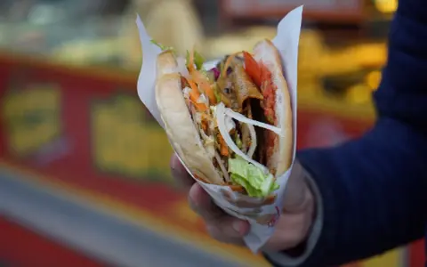 Kebab w bułce z warzywami i mięsem, trzymany w dłoni.