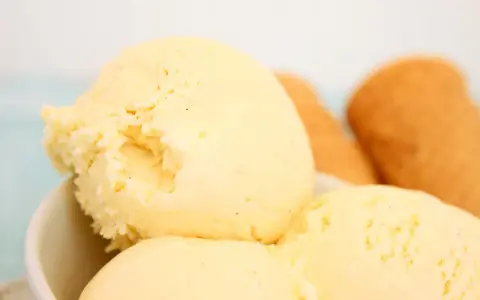 Cremoso helado de vainilla con motas oscuras, servido en un tazón blanco. Al fondo, conos de helado dorados esperan.