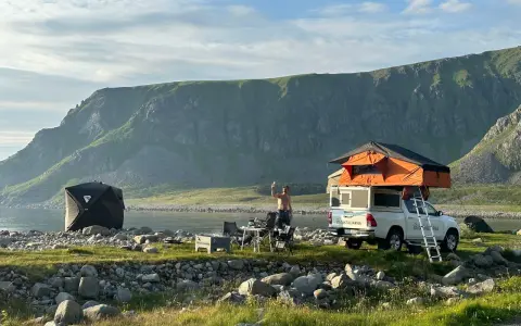 En man tar en selfie vid sitt tak-tält på en strand i Norge. Bredvid står en mörk kupoltält och ett bord med stolar. Berg i bakgrunden.