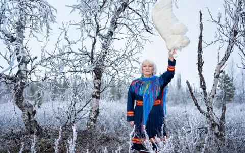 Britta Marakatt Labba i samedräkt håller upp en stor vit fjäder i en snötäckt skog.