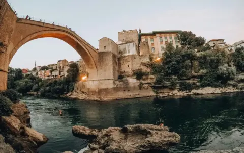 Iconic Stari Most bridge over the Neretva River, a must-see for any Bosnia travel adventure. People watch from the bridge as swimmers enjoy the emerald waters below.