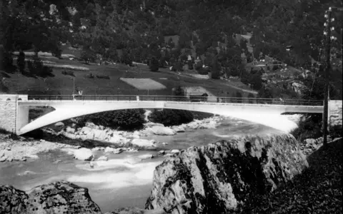 Puente de arco de hormigón de **Robert Maillart** sobre un río rocoso, con colinas boscosas al fondo.