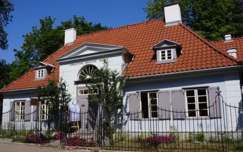 Jasnoniebieski dworek Sierakowskich z czerwoną dachówką, otoczony zielenią i ogrodzeniem.