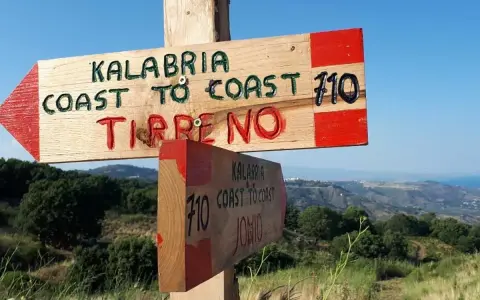 Cartello in legno indica "Kalabria Coast to Coast" verso il Tirreno e lo Jonio, con sfondo di colline e mare.