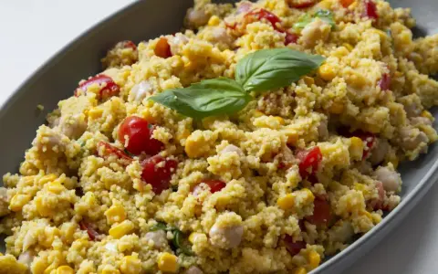 Un piatto colorato con la ricetta cous cous: chicchi dorati, pomodorini, mais e ceci, guarnito con foglie di basilico fresco.