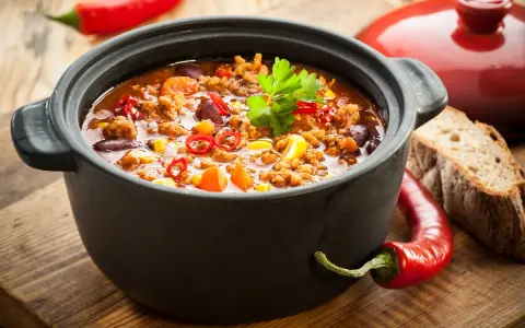 Un plat de chili mijoté dans une cocotte inox four, garni de persil, piments et maïs. Parfait pour un repas réconfortant.