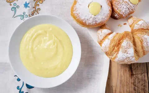 Una ciotola di soffice crema pasticcera senza uova accanto a cornetti e bomboloni spolverati di zucchero a velo.