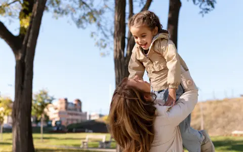 Madre levanta a su hija riendo, un momento lleno de palabras bonitas para niños y amor en un día soleado en el parque.