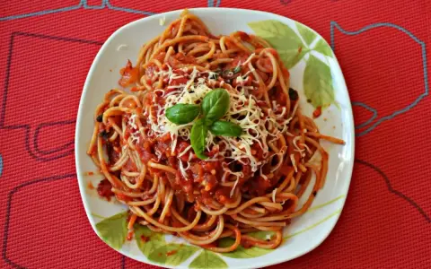 Pyszny sos do makaronu spaghetti, posypany startym serem i udekorowany listkiem bazylii, na talerzu z wzorem liści.