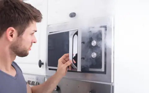 Mann öffnet Mikrowelle, aus der Rauch dringt. Ein Fehler mit Metall in Mikrowelle kann gefährlich sein.