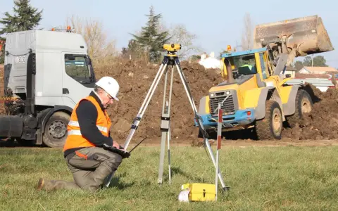 Geodeta w kamizelce odblaskowej nadzoruje odwierty geologiczne pod budowę domu. W tle koparka i ciężarówka.