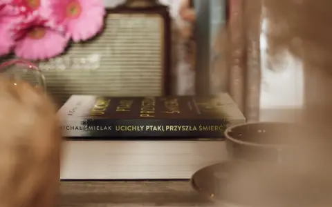 Na stole leżą książki, jedna z nich ma tytuł "Ucichły ptaki, przyszła śmierć". W tle różowe kwiaty i filiżanka.